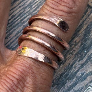 May include: A copper-coloured spiral ring featuring a multi-layered design that wraps around the finger. The ring consists of four distinct bands, with the top and bottom bands displaying a textured, hammered finish, while the two middle bands are smooth.