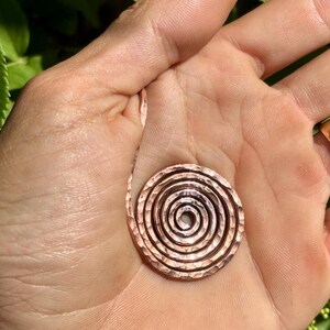 May include: A hammered copper spiral pendant with a textured surface. The pendant is circular, with concentric rings forming a spiral design. The copper has a warm, reddish-brown hue.