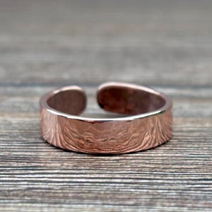 May include: A copper-toned ring with an open band design, featuring a polished, hammered texture that creates a wavy, reflective surface across the exterior of the metal band. The ring has a simple, continuous shape with a gap at the back.