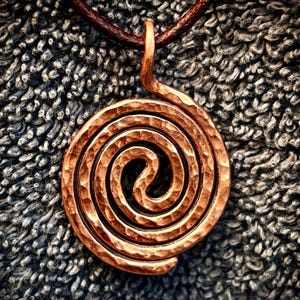 May include: A circular pendant featuring a hammered copper spiral design. The metal surface has a textured, dimpled finish with warm reddish-brown tones. The pendant is suspended from a dark brown leather cord that loops through the top bail.