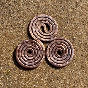 May include: A copper-coloured metal triskelion ornament featuring three interconnected spirals. The surface of the metal displays a distinct hammered texture, creating a series of small, reflective indentations across the entire curved form.