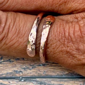 May include: A rose gold-toned bypass ring featuring a hammered, textured surface. The ring consists of two overlapping, curved bands that wrap around the finger, creating a continuous, reflective metallic appearance with irregular indentations.