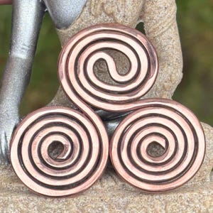May include: A copper-coloured metal pendant featuring a triskelion design composed of three interconnected spirals. The surface of the metal has a polished finish with dark recessed lines that define the spiral patterns, creating a distinct contrast.