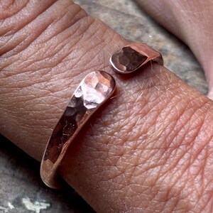 May include: A copper-coloured open ring featuring a hammered, textured surface. The band is crafted with a metallic finish and has two rounded, slightly flattened ends that do not meet, creating a gap in the design of this circular jewellery piece.