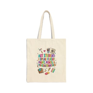 May include: A beige canvas tote bag featuring a colourful graphic print with the text "ART STUDENT: I SPEAK FLUENT PAINT, PENCIL & PROCRASTINATION" surrounded by illustrations of art supplies including pencils, a sketchbook, and paint tubes.