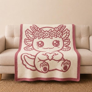Peut inclure: Une couverture en crochet de couleur crème présentant un grand motif rose foncé représentant un axolotl portant une couronne de fleurs et tenant un cœur. La couverture possède une bordure rose foncé unie et un point texturé sur toute la surface.