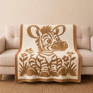 May include: A rectangular crochet blanket featuring a two-tone design in cream and tan. The centre displays a large, stylised zebra sitting among flowers and grass. The blanket is finished with a solid tan border along all four edges.