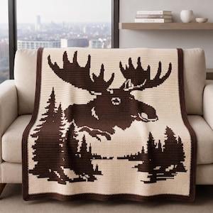 Puede incluir: Una manta de ganchillo rectangular que presenta la silueta de un alce marrón oscuro con grandes astas centrada sobre un paisaje forestal marrón oscuro. La manta está hecha con hilo color crema y tiene un borde marrón oscuro.