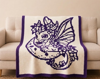 May include: A cream-colored crochet blanket featuring a large, dark purple pixelated graphic of a small dragon with wings and a floral crown, resting on a crescent moon. The blanket is finished with a solid dark purple border on all four sides.