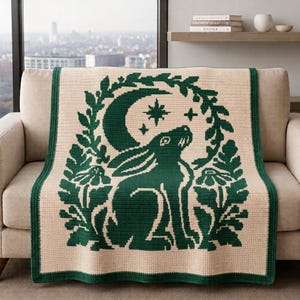 Puede incluir: Una manta de ganchillo rectangular con una base de color crema y un diseño en verde oscuro. El patrón muestra un conejo sentado de perfil, rodeado por una corona circular de hojas y flores, con una luna creciente y estrellas encima.