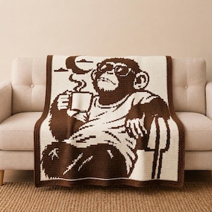 Pode incluir: Uma manta de crochê retangular com um gráfico pixelizado em dois tons, castanho e creme, de um chimpanzé a usar óculos de sol. O chimpanzé segura uma caneca a fumegar, com uma lua crescente e nuvens visíveis no fundo.
