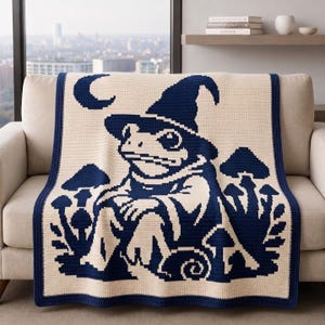 Può includere: Una coperta all'uncinetto color crema con un disegno grafico blu scuro di una rana che indossa un cappello a punta, circondata da vari funghi e una luna crescente. La coperta ha un bordo blu scuro solido che incornicia l'intero pezzo.