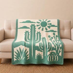 May include: A rectangular crochet blanket featuring a two-tone desert landscape design in cream and sage green. The pattern displays various cacti, a sun, and clouds. The blanket has a solid sage green border framing the entire graphic scene.