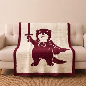 May include: A cream-colored crochet blanket featuring a large, dark maroon graphic of an otter wearing a cape and holding a sword aloft. The blanket has a thick, solid dark maroon border that frames the central design on all four sides.