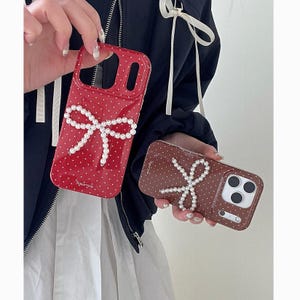 May include: Two phone cases, one red and one brown, each featuring a pearl bow design. The red case has white polka dots. The brown case has white polka dots. Both cases have a glossy finish and are held by a person.