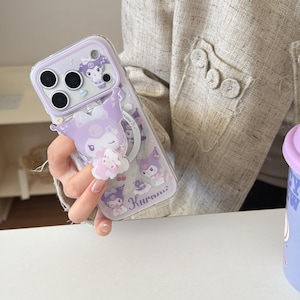 Puede incluir: Una funda transparente para teléfono con un diseño de personaje de dibujos animados morado y blanco. La funda tiene un agarre emergente y presenta el personaje "Kuromi" con estrellas y otros elementos decorativos. La funda está diseñada para un teléfono inteligente.