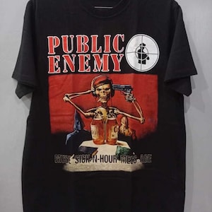 Puede incluir: Camiseta negra con la inscripción roja "PUBLIC ENEMY" y un dibujo de un esqueleto sosteniendo una pistola. La camiseta también tiene el texto "MUSE SICK-N-HOUR MESS AGE".