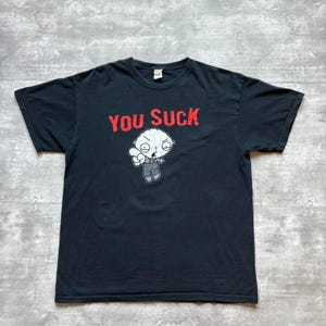 Może przedstawiać: Czarny t-shirt z czerwonym napisem "YOU SUCK" nad postacią z kreskówki. Postać jest biała z zaskoczoną miną i wskazuje palcem. Koszulka leży płasko.