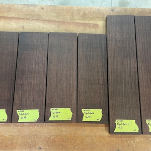 May include: Six rectangular pieces of dark brown wood, possibly wenge, arranged on a light brown surface. Each piece has a yellow sticker with text and measurements. The wood grain is visible.