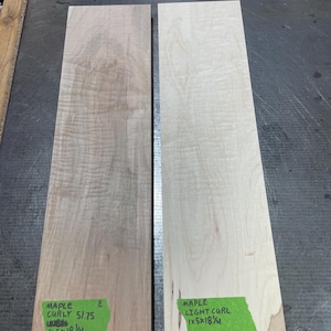 May include: Two rectangular maple wood planks, one with a curly grain and the other with a light curl. The curly maple plank has a sticker that reads "MAPLE CURLY 51.75 1x5x18 1/4". The light curl plank has a sticker that reads "MAPLE LIGHT CURL 1x5x18 1/4".