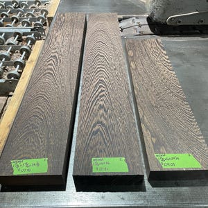 May include: Three rectangular Wenge wood planks with a dark brown and black grain pattern. Each plank has a green sticker with dimensions and price. The planks are 1 1/8" x 5 5/8" x 36 3/8", 1 1/8" x 6" x 36", and 1 1/8" x 6" x 24 1/4".