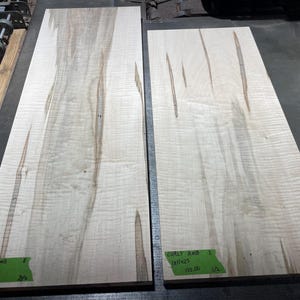 May include: Two rectangular, light-colored wooden boards with a wavy grain pattern. The boards have a natural, light-brown color with darker streaks. Green labels with text are attached to the boards. The boards are likely for woodworking or craft projects.