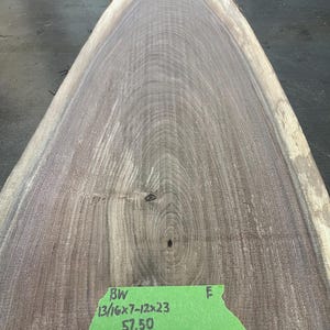 May include: A live edge wood slab with a natural, organic shape. The wood grain features a mix of light and dark brown tones. The slab measures approximately 13/16" x 7"-12" x 23". A green tag displays the dimensions and price.