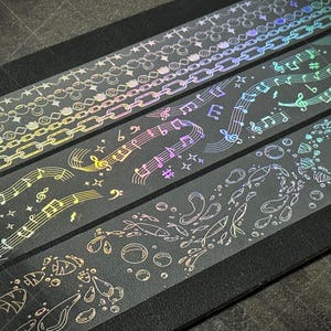 May include: Decorative washi tape rolls with iridescent designs. The rolls feature patterns of chains, musical notes, and fish. The tape is transparent with a holographic effect, creating a shimmering, colorful appearance. Ideal for scrapbooking, journaling, and crafts.