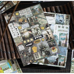 Op de afbeelding: Vellen decoratieve stickers met Japanse tekst en afbeeldingen, waaronder eten, landschappen en stripfiguren. De stickers zijn gerangschikt op een donkere ondergrond met bamboe accenten.