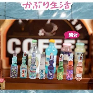 Op de afbeelding: Een verzameling miniatuur Japanse Ramune-sodaflesjes in verschillende kleuren, waaronder blauw, groen en roze. De flesjes staan op een houten plank voor een koffiehuisbord. Japanse tekst is zichtbaar.