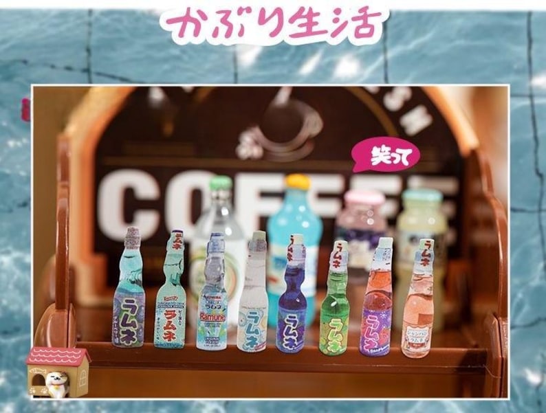 Op de afbeelding: Een verzameling miniatuur Japanse Ramune-sodaflesjes in verschillende kleuren, waaronder blauw, groen en roze. De flesjes staan op een houten plank voor een koffiehuisbord. Japanse tekst is zichtbaar.