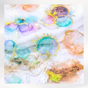 May include: Abstract art piece featuring colorful ink blotches in shades of teal, purple, pink, and gold. The artwork has a watercolor effect with gold glitter accents, creating a dreamy and ethereal aesthetic. The background is a textured white surface.
