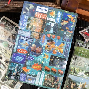 Op de afbeelding: Een vel decoratieve stickers met een blauwe en turquoise achtergrond. De stickers bevatten goudvissen, Japanse tekst, sushi en andere illustraties. Sommige stickers hebben een holografisch, glinsterend effect.