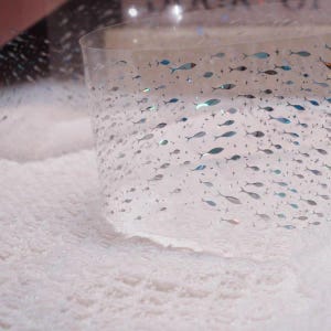 May include: Clear, curved plastic sheet decorated with small, silver and blue fish and star shapes. The sheet is partially resting on a white, fluffy surface. The fish are arranged in a pattern, creating a sense of movement.