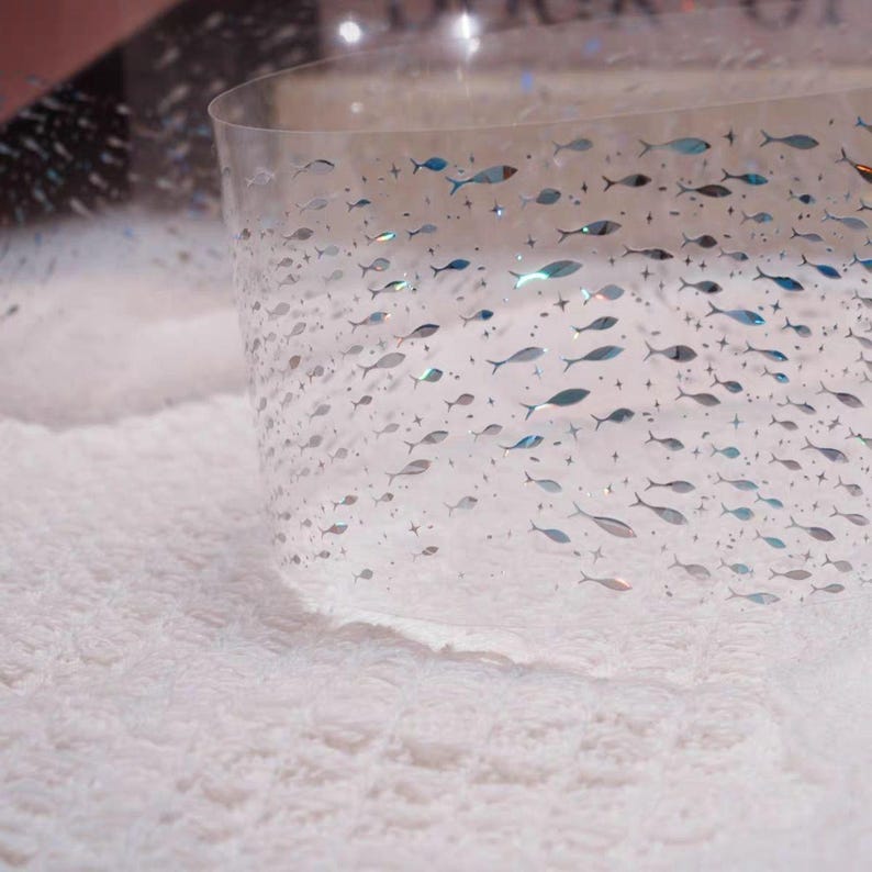 May include: Clear, curved plastic sheet decorated with small, silver and blue fish and star shapes. The sheet is partially resting on a white, fluffy surface. The fish are arranged in a pattern, creating a sense of movement.