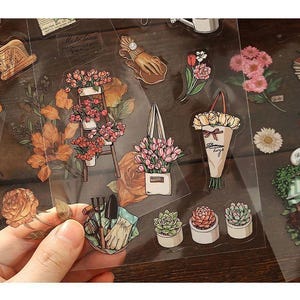 May include: A collection of clear stickers featuring floral and gardening themes. Designs include bouquets, potted plants, gardening tools, and individual flowers in various colors. The stickers are arranged on a wooden surface.