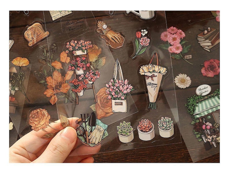 May include: A collection of clear stickers featuring floral and gardening themes. Designs include bouquets, potted plants, gardening tools, and individual flowers in various colors. The stickers are arranged on a wooden surface.