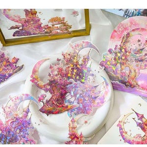 Op de afbeelding: Decoratieve stickers met een fantasiekasteelontwerp in roze, paars en goud. De stickers hebben een holografisch effect en zijn gevormd als manen en kastelen. De tekst "Castle Under" is zichtbaar op een van de stickers.