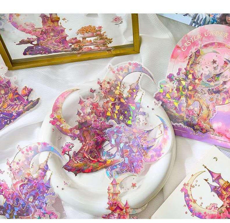 Op de afbeelding: Decoratieve stickers met een fantasiekasteelontwerp in roze, paars en goud. De stickers hebben een holografisch effect en zijn gevormd als manen en kastelen. De tekst "Castle Under" is zichtbaar op een van de stickers.
