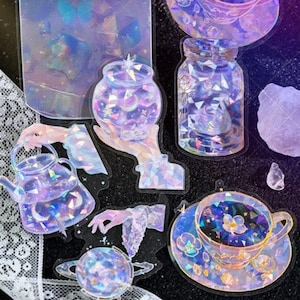 May include: A collection of iridescent stickers featuring celestial and fantasy-themed designs. The stickers include a teapot, teacup, jars, and a globe, all with a sparkling, crystal-like appearance. The color palette is primarily purple, blue, and white.