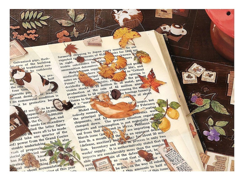 May include: A collection of decorative stickers featuring cats, leaves, butterflies, and fruit, arranged on an open book with printed text. The stickers have a vintage aesthetic with warm colors.