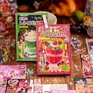 May include: Two sticker packs with Japanese text and illustrations of desserts. One pack is green with a green dessert, and the other is red with a red dessert. A pink clip holds the packs together. Other stickers are scattered around.