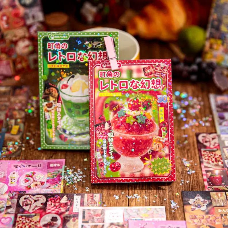 May include: Two sticker packs with Japanese text and illustrations of desserts. One pack is green with a green dessert, and the other is red with a red dessert. A pink clip holds the packs together. Other stickers are scattered around.
