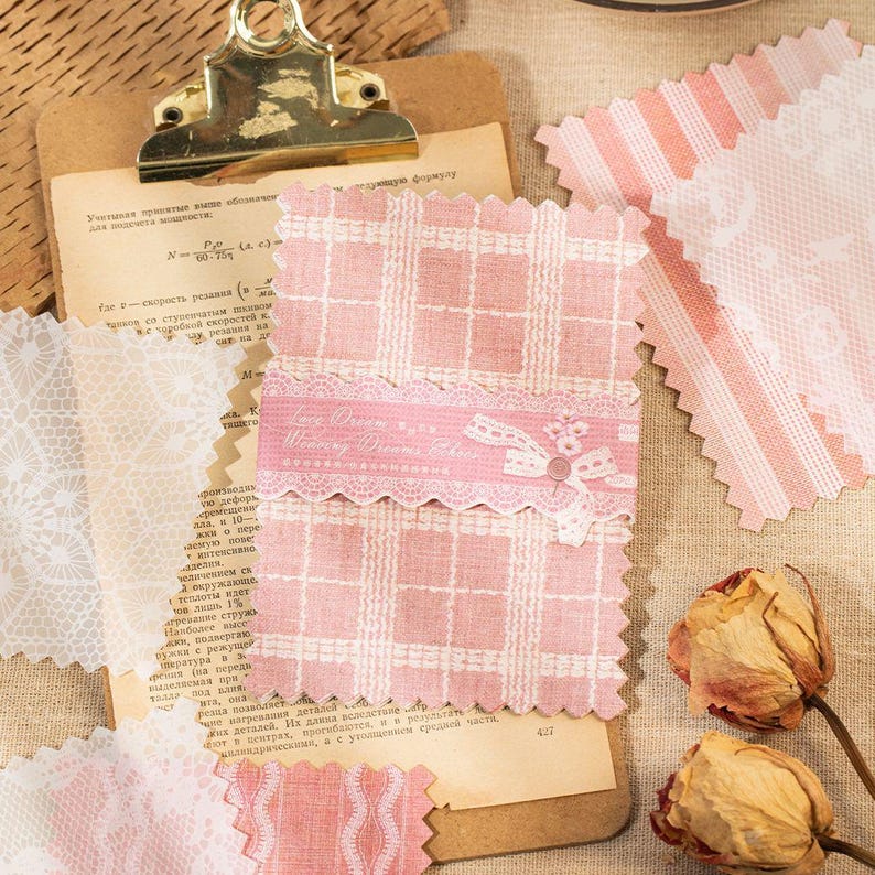 May include: A collection of pink and white patterned fabric swatches, including gingham, lace, and striped designs. A pink ribbon with the text "Winter Dreams Edition" is attached to one swatch. The image also includes dried roses and a clipboard.