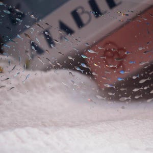 May include: A clear, transparent sheet adorned with numerous small, fish-shaped embellishments. The fish are in various shades of silver and iridescent colors. The background is a soft, blurred white and pink.