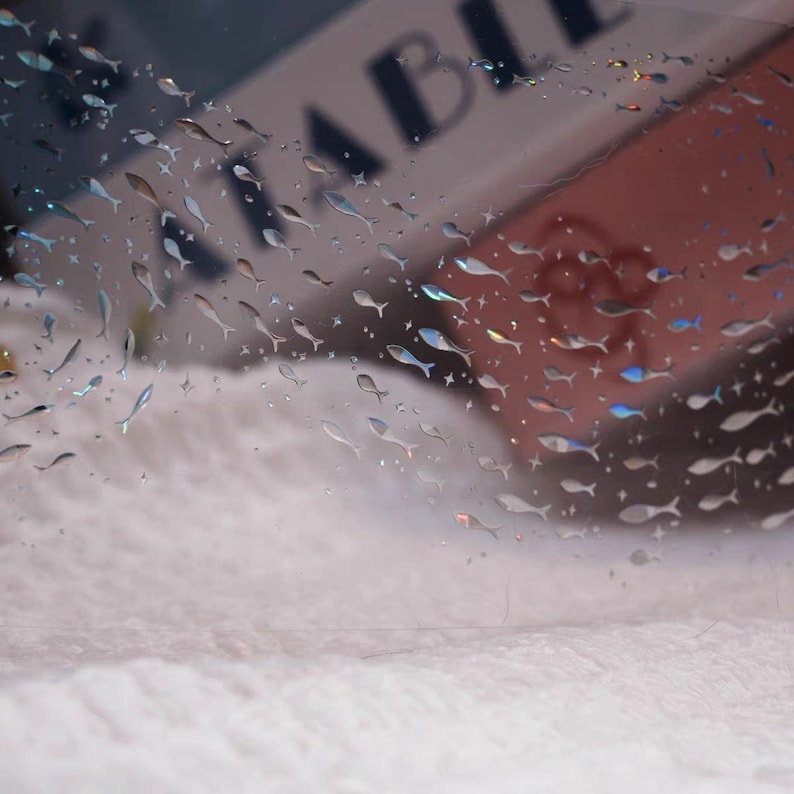 May include: A clear, transparent sheet adorned with numerous small, fish-shaped embellishments. The fish are in various shades of silver and iridescent colors. The background is a soft, blurred white and pink.