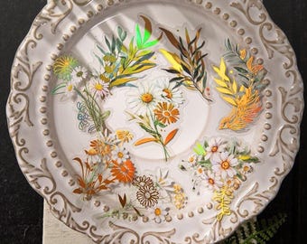 May include: A white, ornate ceramic plate with a raised rim, adorned with floral stickers. The stickers feature daisies, other flowers, and foliage in shades of orange, yellow, and green. The plate has a vintage aesthetic.