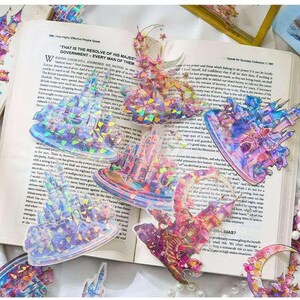 Op de afbeelding: Een verzameling iriserende kasteelvormige stickers met holografische details, verspreid op een open boek. De stickers hebben een reeks kleuren, waaronder roze, blauw en paars, en zijn ontworpen met ingewikkelde architecturale elementen. De pagina's van het boek tonen gedrukte tekst.