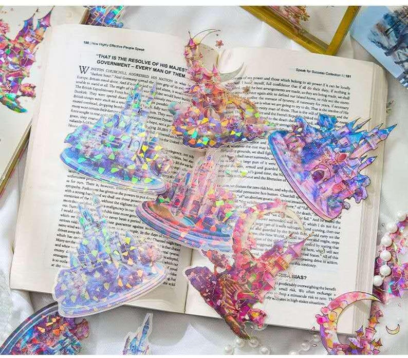 Op de afbeelding: Een verzameling iriserende kasteelvormige stickers met holografische details, verspreid op een open boek. De stickers hebben een reeks kleuren, waaronder roze, blauw en paars, en zijn ontworpen met ingewikkelde architecturale elementen. De pagina's van het boek tonen gedrukte tekst.