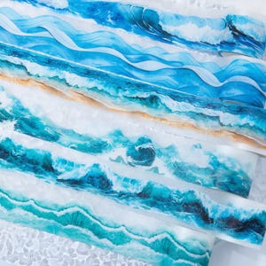 May include: A collection of washi tape rolls with ocean-themed designs. The tape features various shades of blue and white, depicting waves, sandy beaches, and sea foam. The rolls are layered, showcasing the different designs and textures.